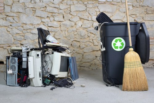 In the foreground, a pile of discarded old computer equipment and electronic waste, including monitors, keyboards, and various peripherals, is placed on a concrete surface. The equipment shows signs of wear, with some cables tangled among the devices. To the right, a large black wheelie bin with a green recycling symbol on the side is positioned against a textured stone wall, with a wooden broom leaning against it. The scene appears to be outdoors, possibly on a driveway or paved area near a residential or commercial property. The overall setting reflects typical rubbish collection for electronic waste and general household refuse, with Woolwich House Clearance providing rubbish removal services in the local area. The natural lighting suggests daylight, emphasizing the mixture of materials and textures present in the waste, ready for disposal or recycling.