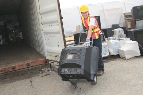 A worker wearing a yellow safety helmet, reflective vest with red and yellow stripes, and work gloves is standing outdoors on a paved surface near an open shipping container. The worker is holding a large, old computer monitor with both hands, positioning it for disposal or removal. Surrounding the worker are various discarded electronic appliances, including multiple beige and white computer towers, CPUs, and other miscellaneous electronic waste items, stacked loosely on the ground and leaning against the shipping container. The background shows the inside of the shipping container, which appears dark and empty, contrasting with the outdoor lighting. This scene depicts a professional rubbish removal associated with waste management services in Woolwich, supporting environmentally responsible disposal of electronic waste, consistent with services offered by Woolwich House Clearance.