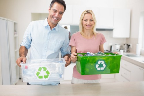 A middle-aged man and woman stand together in a modern kitchen, smiling and holding biodegradable recycling bins with green and white recycling symbols, ready for waste sorting. The man, dressed in a light blue shirt, holds a transparent bin filled with plastic bottles, while the woman, wearing a pink top, carries a solid green bin. The background features a white kitchen with sleek cabinetry, a countertop, and various utensils, indicating a clean and organized environment. The scene is well-lit with natural or ambient lighting, and the focus is on the individuals and their recycling containers, emphasizing responsible waste separation practices commonly associated with rubbish removal services in Woolwich and surrounding areas. Woolwich House Clearance often assists with clearing household waste, including recyclable materials, in the local community.