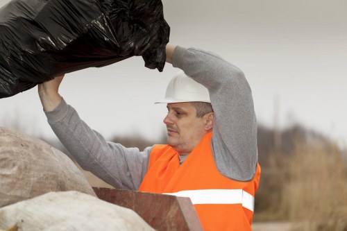 A male waste collection worker wearing a white hard hat and an orange high-visibility vest is outdoors, lifting a large black plastic rubbish bag above his head to load it into a container or skip, which is partially visible in the foreground. The background shows a muted, overcast sky and a natural landscape with some blurred vegetation, suggesting a rural or suburban setting near Woolwich. The worker appears focused on his task in a professional manner typical of rubbish removal services provided by Woolwich House Clearance. The scene emphasizes the manual handling of waste materials, with additional rubble or debris visible on the surface next to him, and the environment is free of clutter or immediate obstacles, indicating an organized clearance operation.