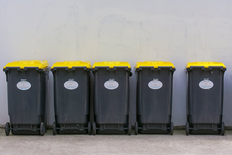 A black wheeled rubbish bin with a white label and the text 'ST. JOHN'S' printed on the front, positioned on a paved sidewalk adjacent to a curb. The bin is cluttered with assorted waste items, includ
