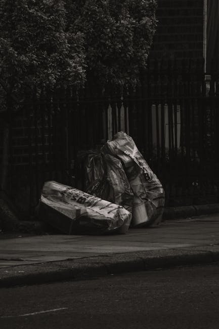 Three large black garbage bags, made of durable plastic with crinkled and slightly shiny surfaces, are stacked against a black metal fence on a paved sidewalk. The bags appear to contain household was